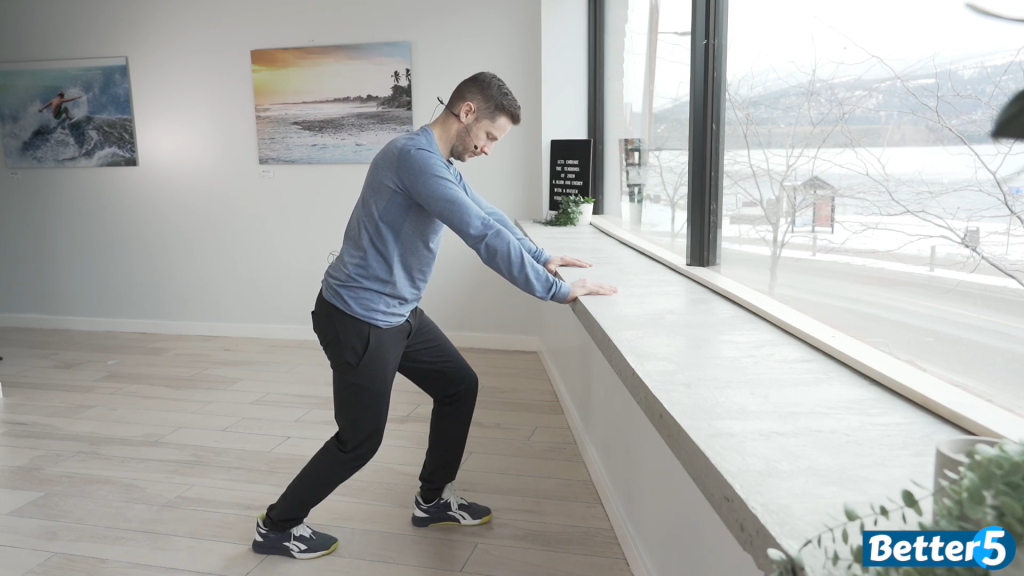 Senior performing a calf stretch exercise by leaning on a counter, extending one leg back to stretch the calf muscles, as part of the 10 exercises to prevent falls.
