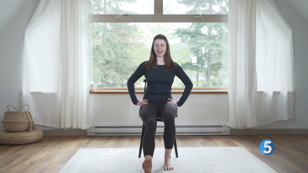 A fitness instructor is seated on a chair in a bright room with large windows. She is performing a heel drag exercise. This exercise helps in enhancing leg strength and flexibility, making it ideal for seniors.
