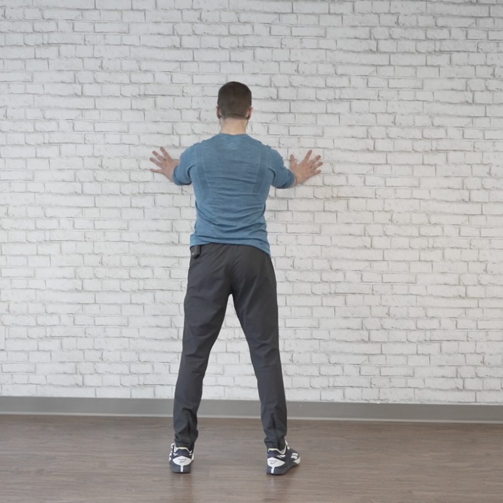 Fitness instructor demonstrates how to perform wall push-ups, a simple arm exercise for seniors targeting arm & shoulder muscles.