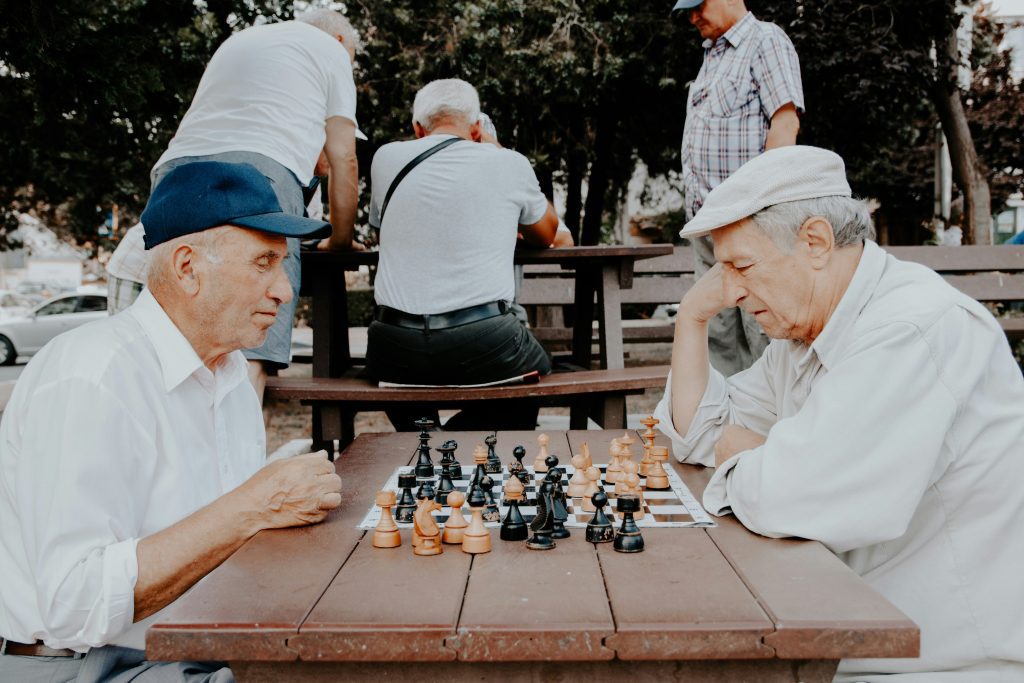 Two senior men sitting at a wooden table outdoors, deeply focused on a game of chess, a popular memory game for seniors that enhances strategic thinking and mental sharpness.
