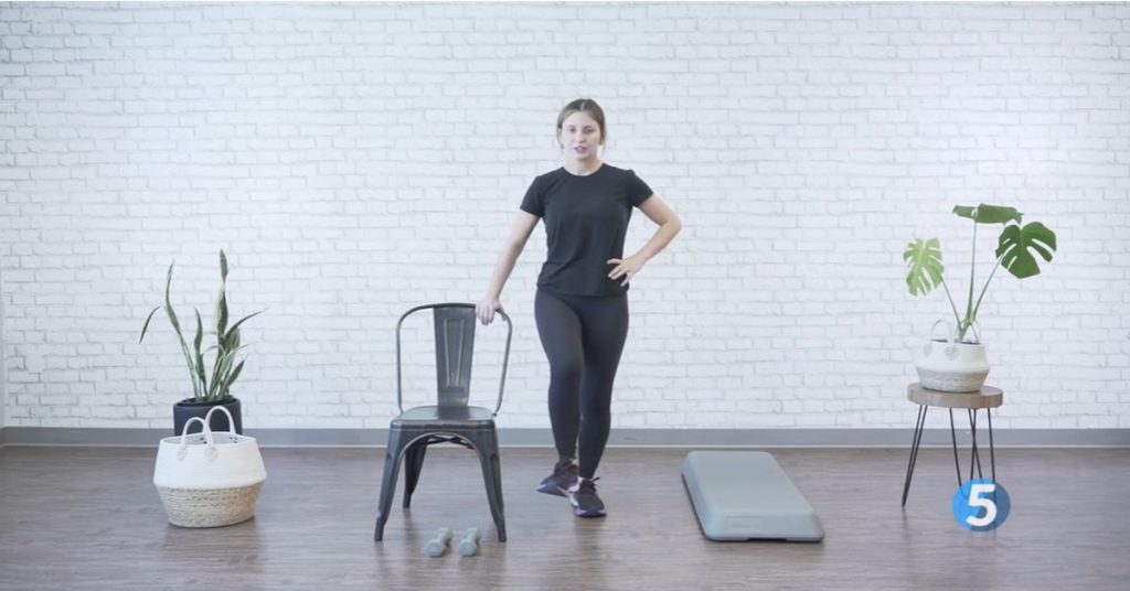 Instructor demonstrating a standing march core exercise for seniors, lightly holding a chair for balance support