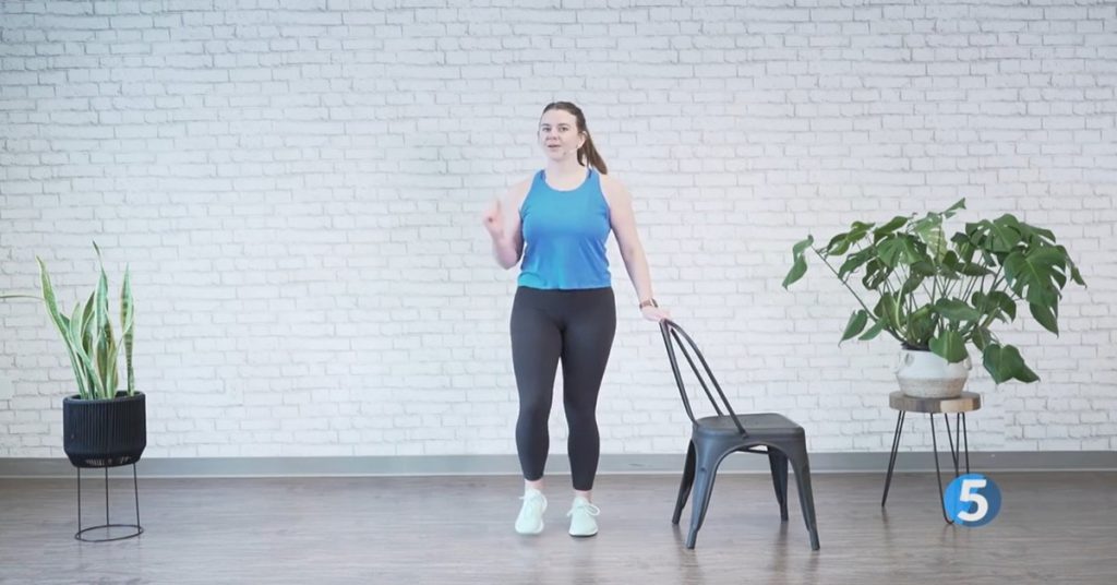 female instructor demonstrating a chair-supported standing march for seniors