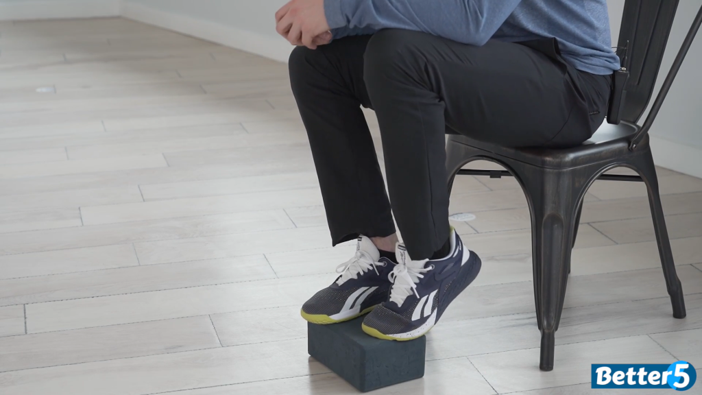 The fitness coach demonstrates a great ankle mobility exercise by placing the ball of his foot on the yoga block and raising his heel up.