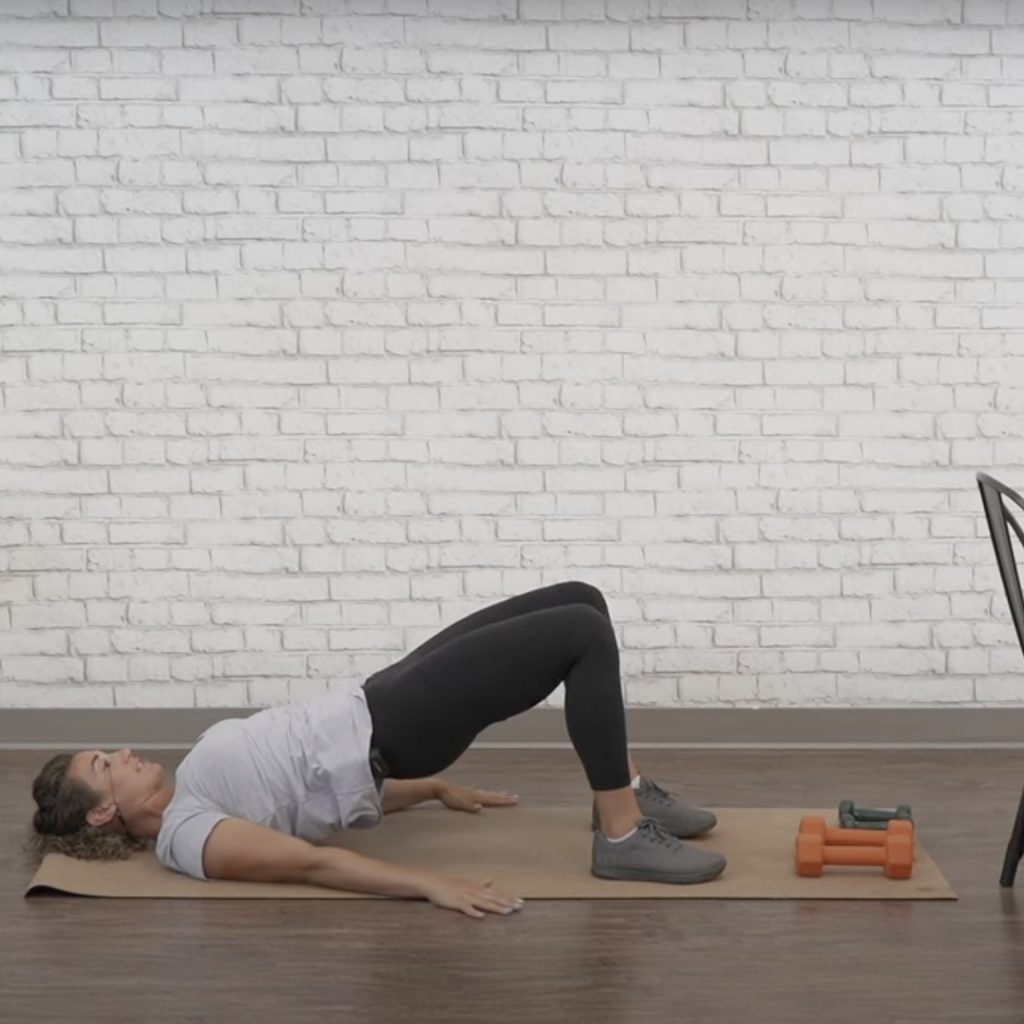 The fitness instructor demonstrates the bridge position as part of pelvic floor therapy at home, lying on a mat with her hips lifted and feet on the ground.