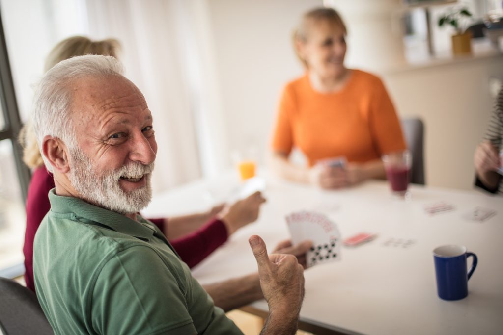 Senior man leans back confidently smiling, as he plays cards with close friends–a great way to improve mental health in older adults.