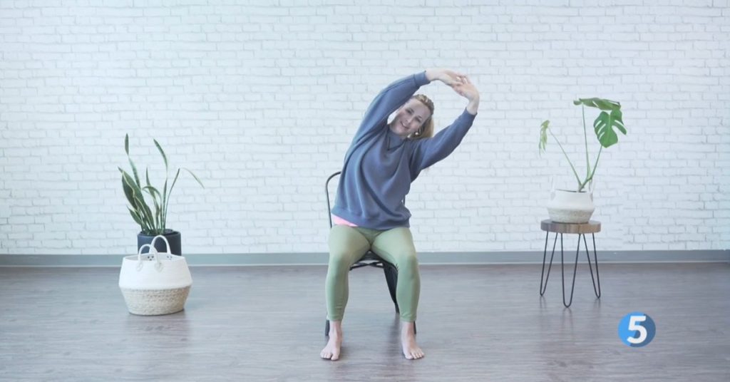 Female instructor performing seated chair yoga side bend for seniors, gentle at-home stretch to improve flexibility and posture.