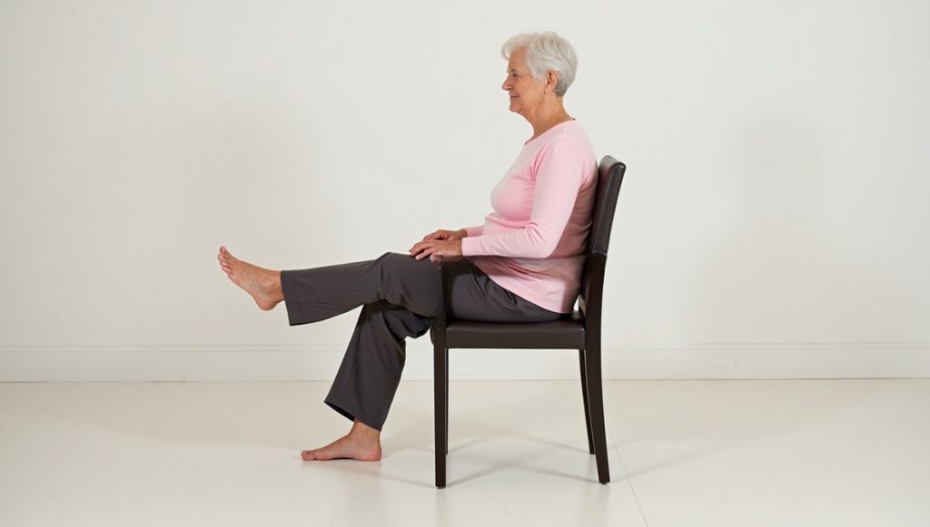 A senior woman sitting indoors demonstrates how to perform a seated knee extension.