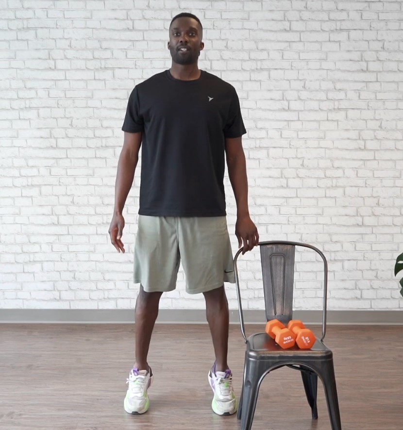 A fitness instructor in a black t-shirt and gray shorts performs calf raises near a chair with orange dumbbells on the seat.
