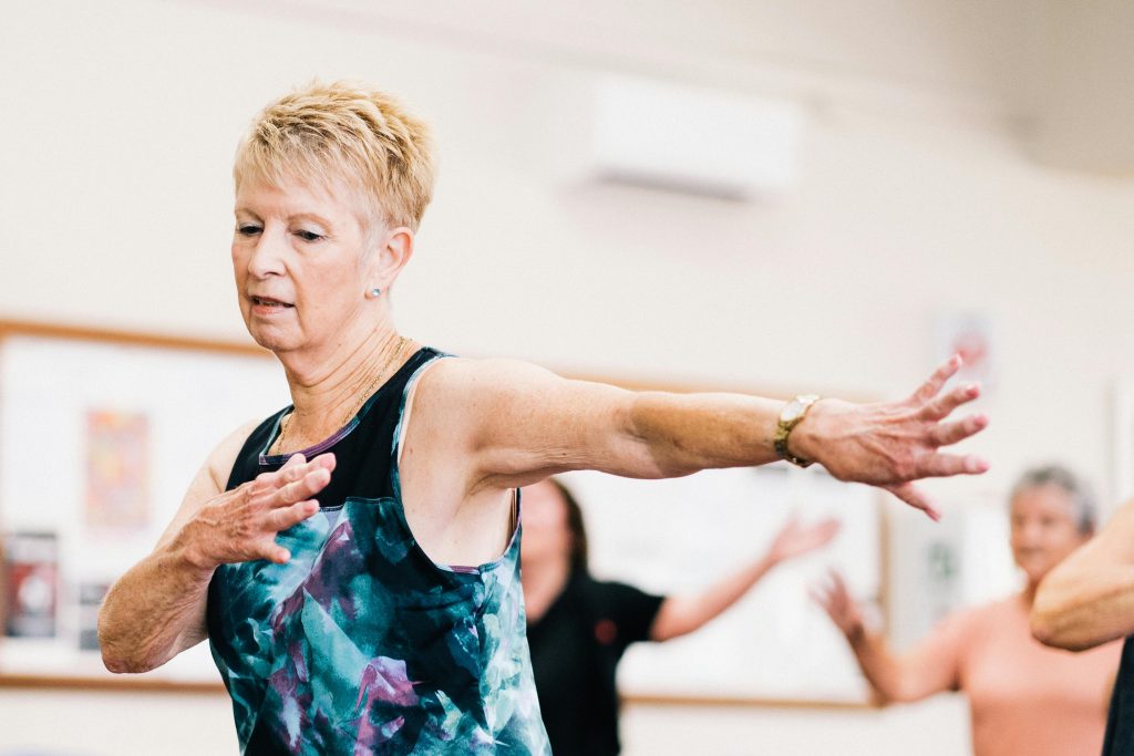 An elderly woman extends her arms in an arm stretch during a fitness class, highlighting why it's never too late to start exercising for strength and flexibility.