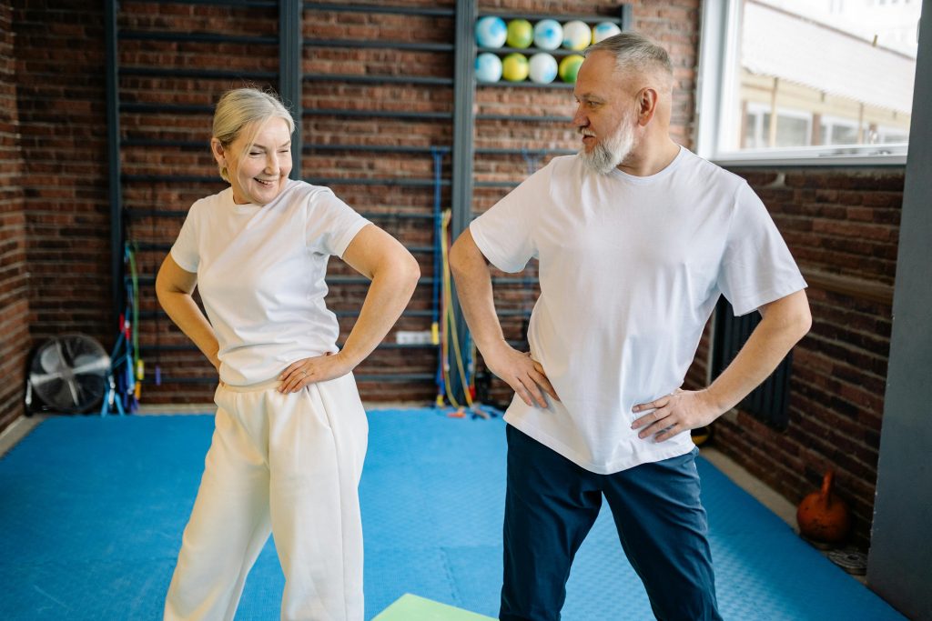 smiling elderly couple in a gym setting stands with hands on hips, engaging in a warm-up routine.