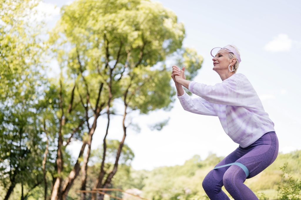 An elderly woman performs resistance band squats, demonstrating why it's never too late to start exercising and build strength at any age.